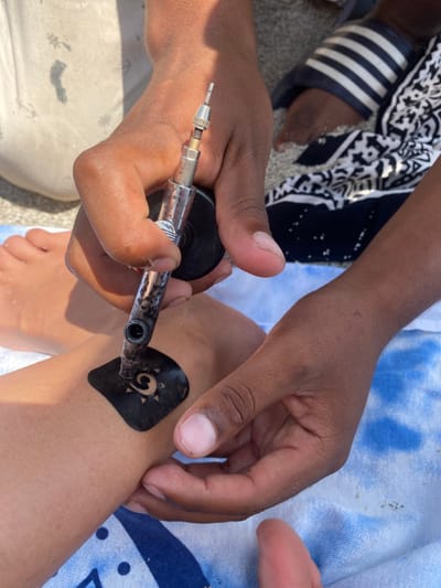 A person is getting a henna tattoo applied to their ankle by another person, possibly a professional henna artist. The artist is holding a metal henna applicator with a black nozzle and applying it to a black stencil on the ankle. The person getting the tattoo is sitting on a blue and white beach towel. The location is in Punta Hermosa, Peru, which is a district in the province of Lima, with the coordinates -12.326 latitude and -76.835 longitude. The scene is on a beach or near the beach as sand is visible and a striped towel is in the background. The artist's other hand is holding the stencil in place. The artist is wearing a black and white patterned shirt with a black belt. The person receiving the tattoo is wearing a white shirt. The environment is sunny and the lighting is bright.