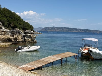 A tranquil bay in Kassopaia Municipal Unit, Greece, is captured under a clear blue sky. The scene features a small, pebble beach leading to crystal-clear water where a wooden pier extends into the sea. Two inflatable boats are anchored nearby. The larger boat, a white rigid-hulled inflatable with an outboard motor, is positioned closer to the shore. Further out, a white motorboat with a canopy is moored. In the background, rolling green hills and rocky outcrops define the coastline, with the vast expanse of the sea stretching towards a distant hazy horizon. A sailboat is visible in the distance, adding to the serene maritime atmosphere. The water is calm, reflecting the bright sunlight and showcasing the submerged pebbles beneath the surface. The overall impression is one of peaceful seclusion and natural beauty.