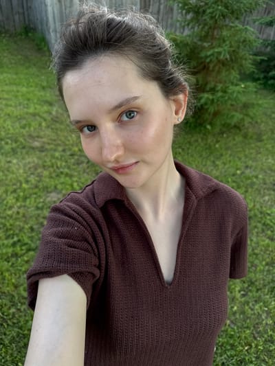 A young woman with fair skin, light blue eyes, and dark brown hair pulled up is shown in a selfie. She is wearing a brown knit shirt with a collar and a deep V-neck. The background consists of a wooden fence, green grass, and a tree. The lighting suggests it is daytime, possibly outdoors. The location is the city of Burdyata, Russia.