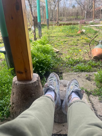 The scene depicts a first-person view of a person's lower legs and feet, extending outward into a garden or yard in Blaskovo, Bulgaria. The individual is wearing light green trousers with ribbed cuffs and white athletic socks, paired with light grey, mint green, and light brown athletic shoes featuring pink accents. Their feet rest on a light-colored concrete step or natural stone.

To the left, a substantial square-cut wooden post, exhibiting natural wood grain and knots, stands vertically, supported by a rough, rounded concrete or stone base. Adjacent to the wooden post on the left is a dark green vertical pole with a black base. Further left, a small black solar light is visible.

The immediate foreground around the steps and bases features lush green foliage. Beyond the steps, a grassy area dominates the midground, interspersed with numerous yellow dandelions and other green plants. Towards the middle-right, a terracotta-colored, bird-shaped planter contains a cluster of artificial or real light-colored flowers, primarily white and pink. Further to the right, a large earthenware jug or pot with a wide neck is partially visible. Concrete stepping stones are visible in the grassy area leading away from the viewer.

The background consists of a rustic outdoor environment with several bare deciduous trees and shrubs, indicating early spring. Various green-painted metal posts and wire fencing are visible, defining the perimeter or sections of the garden. A light blue sky with some cloud cover is visible above the tree line.