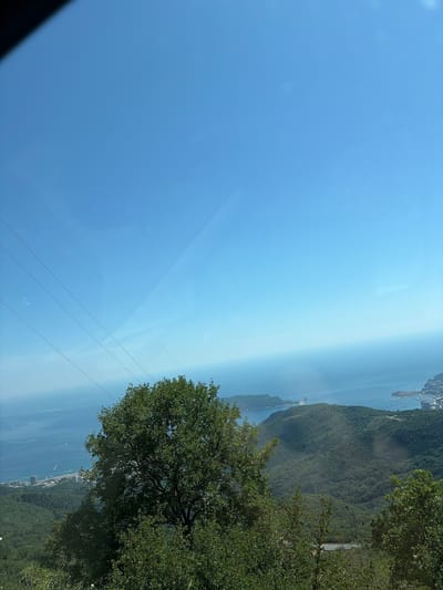 The image captures a scenic view from a high vantage point overlooking the sea and a city in Stanišići, Montenegro. In the foreground, a large, leafy green tree dominates the right side of the frame, its branches reaching upwards. Below and to the left, dense green foliage and trees cover the hillside. The middle ground reveals a lush, green mountainous landscape with a winding road visible in the distance. The background showcases the vast, blue expanse of the sea, with a coastline dotted with buildings and boats. The sky above is a clear, bright blue. Four parallel power lines stretch diagonally across the upper left portion of the image. The overall impression is a serene and picturesque coastal landscape.