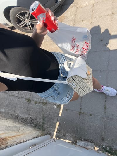 This eye-level, top-down perspective photograph captures the lower body of a person standing outdoors on a sunny day in Rakovski, Bulgaria. The person is wearing distressed light-wash denim shorts, a black top or bodysuit, and white athletic shoes with subtle pink accents. They carry a white woven straw handbag with a matching shoulder strap, slung across their body.
In their left hand, the person holds a red Red Bull energy drink can, which appears slightly dented. The can features the brand's distinctive white bull logo. Resting on the ground near their left foot is a transparent white plastic shopping bag with red Cyrillic text. The text, which is partially obscured and mirrored due to the bag's material, appears to include words like "МЛЯКО" (milk) and "ДЕЛИКАТЕСИ" (delicacies), suggesting grocery items. The person's nails are visible, painted with a light color and a delicate design.
The ground consists of grey concrete pavers with some visible cracks, weeds, and debris. Strong sunlight casts sharp shadows, indicating a bright, clear day, likely midday or early afternoon. To the person's left, part of a white car, including a wheel with black and silver spokes, is visible. On the right side of the frame, a concrete curb and a light grey metallic structure or wall suggest an adjacent building or fence. The scene depicts a casual moment, possibly after shopping, in an urban or semi-urban setting.