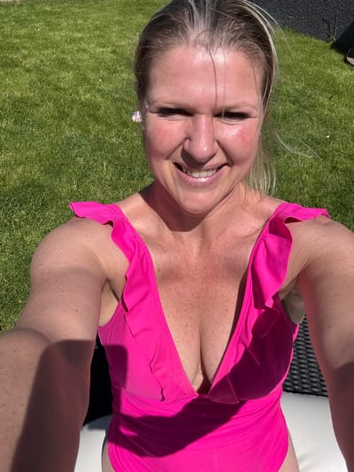 An adult woman is captured in a close-up, outdoor selfie, smiling broadly at the camera. She has blonde hair, with some strands appearing windblown around her face. Her eyes are partially closed in a joyful expression, and she displays a wide, toothy smile, revealing white teeth and a glimmer on her lips. Her skin appears fair with some rosy tones on her cheeks and nose, possibly from sun exposure or a healthy flush. She is wearing a vibrant, fuchsia-pink swimsuit, characterized by ruffled detailing along the neckline and straps, which are visible across her shoulders and décolletage. A small, colorful earring can be faintly seen in her right ear.
The woman is reclining outdoors, likely on a sun lounger, with a dark, possibly wicker-patterned surface visible behind her left shoulder and upper back. A light-colored surface, possibly a cushion or towel, is visible on her right side. The background consists predominantly of lush green grass, suggesting a lawn or garden setting. A dark grey or black paved area is visible in the upper left corner of the frame.
The scene is bathed in bright sunshine, casting distinct shadows across her body and the immediate surroundings, indicating a clear, sunny day, likely midday or early afternoon. The overall mood is one of happiness and relaxation, as conveyed by her cheerful expression. The photo is set in an outdoor location, specifically noted as Marche-en-Famenne, Belgium. No visible text is present in the image.