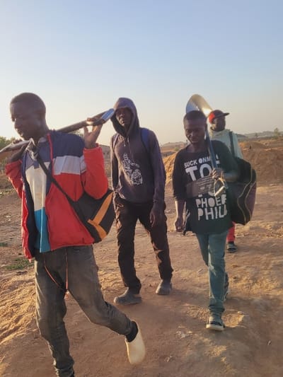 An outdoor scene in what appears to be an undeveloped area of the city of Shen, Nigeria, depicts four males walking on an unpaved dirt path under a clear, light blue sky. The ground consists of uneven, sandy terrain with small mounds. In the mid-ground, large piles of sand or excavated earth are visible, suggesting construction or quarrying activity.
From left to right:
1. A young adult male, dark-skinned, moves towards the left, carrying a shovel over his right shoulder. He wears a red, white, and blue color-blocked jacket, dark grey jeans, and a single white clog on his right foot. A black and orange shoulder bag is slung across his chest.
2. A young adult male, dark-skinned, walks behind the first, looking forward. He wears a dark grey hooded sweatshirt with white graphics, dark pants, and dark grey patterned slip-on shoes, with a dark backpack.
3. A young adult male, dark-skinned, walks to the right of the second, looking downwards. He carries a large brass instrument bell, consistent with a sousaphone or tuba, on his left shoulder and a dark duffel bag in his right hand. He wears a dark green long-sleeved t-shirt with white text and light blue jeans with dark sneakers.
4. Partially visible in the background, an older male, dark-skinned, walks towards the right. He wears a light green/khaki top and a red baseball cap.
The scene is illuminated by natural daylight, casting soft shadows, consistent with an outdoor setting during daytime. No direct interaction between the individuals is observed, though their proximity suggests they are a cohesive group.