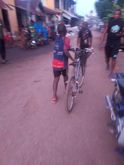 The image depicts a street scene in Orlu, Nigeria. A young boy is walking with a bicycle. Two other individuals are seen walking on the street in the background. There are buildings visible along the street, some with stalls or displays outside. A motorcycle is parked on the side of the road. The road surface appears to be reddish.