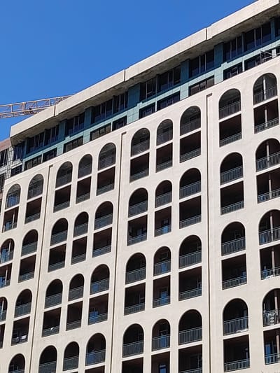 The image shows a modern, multi-story building under construction in Makhinjauri, Georgia. The building features a beige facade with numerous arched balconies, each equipped with dark metal railings. Above the beige section, there is a level with large windows and a teal-colored accent. Further up, the building continues with darker windows and exposed construction elements, including a yellow construction crane visible in the upper left corner against a clear blue sky. The scene depicts urban development in a coastal city.