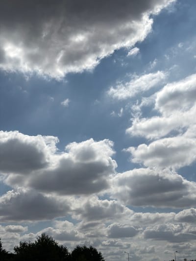 The image depicts a sky filled with a variety of clouds, ranging from dark, heavy formations to lighter, wispy ones, against a backdrop of blue sky. The clouds are illuminated by sunlight, creating contrasts of light and shadow. In the lower portion of the image, the dark silhouettes of trees and vegetation are visible, suggesting an outdoor setting. There are also two streetlights visible in the distance. The overall scene is one of a cloudy sky above a natural landscape. There is no indication of specific urban infrastructure, human activity, or any discernible landmarks that would pinpoint the location to Évry-Courcouronnes, France, beyond it being an outdoor, presumably somewhat populated area given the streetlights.