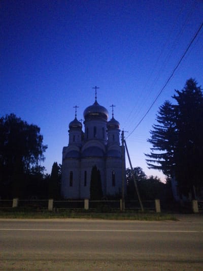 This media file depicts a religious building, likely a church, silhouetted against a deep blue twilight sky. The building features multiple domes topped with crosses, with the largest central dome being gilded. The architecture appears to be Eastern Orthodox, characterized by its rounded domes and crosses. The church is partially obscured by trees on the left and a large pine tree on the right. In the foreground, a paved road runs horizontally across the frame, with a white line marking the center. A low fence with white posts separates the road from the church grounds. Electrical wires and a utility pole are visible in the sky, suggesting the presence of civilization. The overall mood is serene and peaceful, with the fading light of dusk lending a sense of quietude. The time of day is clearly evening, as indicated by the deep blue color of the sky transitioning towards night. There is no visible text in the image. The context provided suggests this scene is located in Lanivtsi, Ukraine.