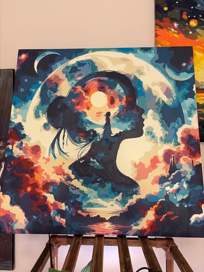 The image shows a square canvas painting. The painting depicts a silhouette of a person or figure floating within a celestial scene. A crescent moon and full moon are present. Colors include dark blues, reds, oranges, and lighter creams. The style is somewhat abstract. The canvas is mounted on a wooden easel or display stand. Another painting is partially visible to the left, and a section of a painting is partially visible in the foreground, just below the main painting. It is unknown from the image whether the painting is located in Woodstock, Canada.