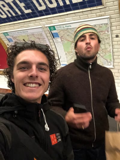 Two adult males are depicted in a selfie-style photograph taken within what appears to be the Porte de Charenton Métro station in or bordering Charenton-le-Pont, France. The background consists of white tiled walls, with blue tiled signage above displaying partial white lettering "PORTE DU". Two framed maps are mounted on the wall; the left map shows "Val-de-Marne" and route lines, while the right map clearly indicates "Paris" with Métro and information symbols.
The individual in the foreground, presumed to be taking the photograph, has fair skin, dark curly hair, and blue eyes. He is smiling, revealing upper teeth, and looking directly into the camera. He wears a black hooded jacket with a white drawstring, a black shoulder bag strap, and white wired earphones hanging from his neck. Sunglasses are perched on his head, resting in his hair. A patch with a red background and white/orange text/logo is affixed to his right chest.
The second individual, positioned behind and to the right, has fair skin, dark facial hair, and dark eyes. He wears a multi-colored (green, yellow, red, brown) striped knit beanie and a dark brown zippered turtleneck jacket. He holds a dark rectangular object, possibly a mobile phone, in his left hand, which features a gold ring on the ring finger. His right hand holds a light brown paper bag. His lips are visibly pursed in a distinct pouting expression.