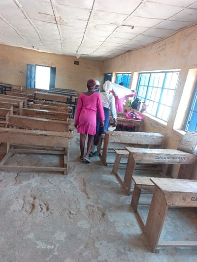 The image depicts the interior of a worn classroom located in Mada, Nigeria. The room features light brown, textured walls, heavily marked with white inscriptions and graffiti. An off-white, grid-patterned ceiling shows signs of damage and discoloration, with visible brown stains and missing panels. The floor is a light brown, uneven concrete surface, displaying extensive flaking and dust.

Natural light enters the room through a series of rectangular windows with blue frames and gridded panes along the right wall. An open doorway with a blue frame is visible on the far left wall, leading to a brightly lit exterior.

The room contains multiple rows of old, light-colored wooden desk-and-bench units. These units are simple in construction and appear worn, with some displaying dark inscriptions such as "CLAP JHUN ACHIEVE CUC SSB" and "HENRY ADAGARY".

Four individuals are present in the middle-right section of the classroom. One person, seen from the back, wears a solid pink dress and light-colored flat shoes, facing towards the right side of the room. Adjacent to this individual, another person wears a white long-sleeved top, a blue skirt, and dark shoes, partially obscured. Further right, near the windows, a person in a pink top and white head covering appears to be handling a white cloth or item. A fourth person, largely obscured, stands nearby, also wearing a white head covering and a white top. A black backpack rests on a desk near the window, and another bag resembling an "iPhone" branded item is on a desk in the foreground right.