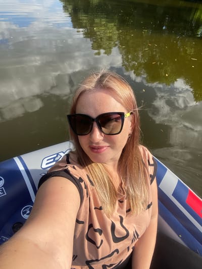 This is a portrait photograph taken from a high angle of a young woman with blonde hair and freckles, wearing sunglasses and a light brown t-shirt with a black abstract design. She is lying on her back on an inflatable object, possibly a paddleboard or an air mattress, which is on a body of water. The water is dark green and reflects the sky, which is partly cloudy with white clouds. Trees are reflected in the water on the left side of the image, suggesting a natural setting. The woman appears relaxed and is looking directly at the camera with a slight smile. The sun is shining brightly, indicating it is daytime, likely a sunny day. The inflatable object has blue and white stripes and some visible text in white, including what looks like a logo or brand name with letters and numbers. One visible instruction or label on the inflatable reads "0.25 MPA 1.5PSI". There are some small dark specks on the surface of the water, possibly leaves or debris. The overall mood of the image is peaceful and summery. The location context is Chulki-Sokolovo, Russia.