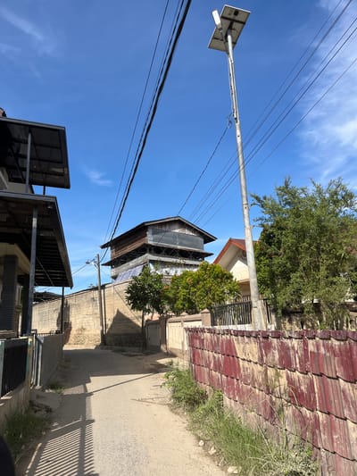 The image depicts a narrow, unpaved alleyway in a residential area of Kota Kuala Simpang, Indonesia. The alley is flanked by a tall, rough-textured concrete wall on the left and a red, corrugated metal fence on the right. The wall on the left has a black metal gate visible in the foreground. On the left side of the alley, a modern house with a dark metal roof structure extends into the frame.

Dominating the sky in the upper right quadrant is a tall metal pole supporting a solar panel and a street lamp. Several electrical wires crisscross the sky, strung between poles not fully visible in the frame. In the background, the alley opens up to reveal more houses with tiled roofs and lush green trees. A distinctive multi-story wooden structure with exposed beams and a dark roof stands prominently in the middle distance, partially obscured by trees. The ground of the alley is dirt and gravel, with some sparse green vegetation growing along the edges. The sky is a clear blue with scattered white clouds. The overall impression is one of a sunlit, somewhat cluttered urban environment.