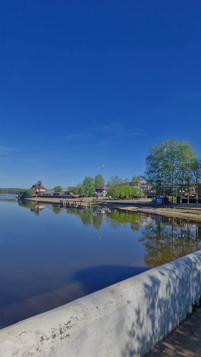 The image shows a canal in Otchor, Russia, on a sunny day with a clear blue sky. Along the canal bank, there's a path lined with trees and streetlights. Buildings are visible along the path, reflected in the still water of the canal. A large concrete barrier is present on the right side of the image, casting shadows on the water. The perspective is unusual, giving the impression that the sky is the water and vice versa.