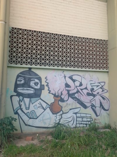 In Cuiabá, Brazil, this image captures a section of an outdoor wall adorned with graffiti. The wall is divided into three horizontal bands. The uppermost section consists of white brickwork. Below this is a rectangular lattice made of repeating circular openings, providing a visual screen. The lower portion of the wall is painted a light yellow-green and serves as the canvas for graffiti art.

The graffiti depicts two figures. On the left is a character with a dark, mask-like face, large eyes, and a wide, toothed grin. It wears a collared shirt. This character is holding a brown object, possibly a vase or jug, from which stylized pink and white smoke or flames emanate. To the right of this figure is a spray-painted word in a graffiti style, rendered in pale pink and outlined in black. The word is complex and stylized, typical of tag art. Below the graffiti, the ground is covered with patchy green grass and weeds. A small red brick lies on the ground near the bottom left of the image. A vertical pipe runs along the left edge of the wall. The overall scene suggests an urban environment where public art has been applied to a building's exterior.