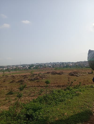 The image shows an outdoor panoramic view of a semi-urban area. In the foreground, there is a patch of dry, brown earth with some sparse green vegetation, rocks, and what appear to be discarded branches or debris. Moving towards the middle ground, there is more cultivated land with rows of crops, and scattered trees. Further back, a densely populated residential area stretches across a gently sloping hill. The buildings are a mix of single-story and multi-story structures with red-tiled or corrugated metal roofs. Several communication towers are visible above the rooftops. The background consists of a hazy, light blue sky with a few wispy clouds. The overall impression is of a developing settlement on the outskirts of a larger town or city. Given the style of buildings and the vegetation, this scene is consistent with the outskirts of Tasha, Nigeria. On the far right of the image, a large, dark, tarp-covered object is partially visible, suggesting some sort of cargo or covering.