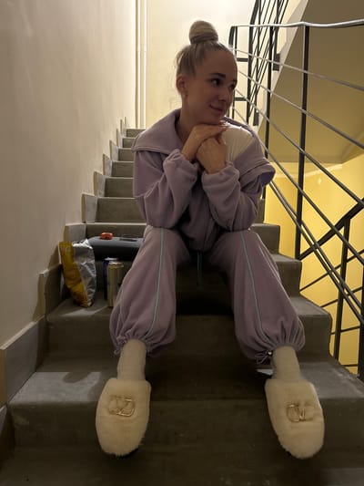 A Caucasian woman with fair skin and blonde hair pulled back into a bun sits sideways on a concrete staircase, her head turned to the left. She wears a light purple tracksuit with turquoise trim and large, furry white slippers with gold embellishments. Her hands are clasped together under her chin.
To the left of the woman are beige walls and black metal stair railings. A bag of chips, a can of something, and a black object rest on the steps near her.
The photo was taken in what looks like an apartment building. The stairs are in Akulovo (Gp Kubinka), Russia.