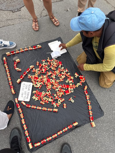 The image depicts an overhead shot of a scene likely taking place on a street in Pisa, Italy. A man with a blue cap marked "Italia" is kneeling near a black cloth spread out on the gray asphalt. The cloth displays a collection of wooden letters and trains, designed to spell out names. A sign nearby indicates "YOUR NAME ONE LETTER €0,60 CENT", suggesting the man sells personalized names. Several people stand around the setup, likely customers, as indicated by their presence nearby. The environment includes surrounding street features like a manhole cover labeled “REXEL ACQUEDOTTO SAINT-GOBAIN CONDUITE”.