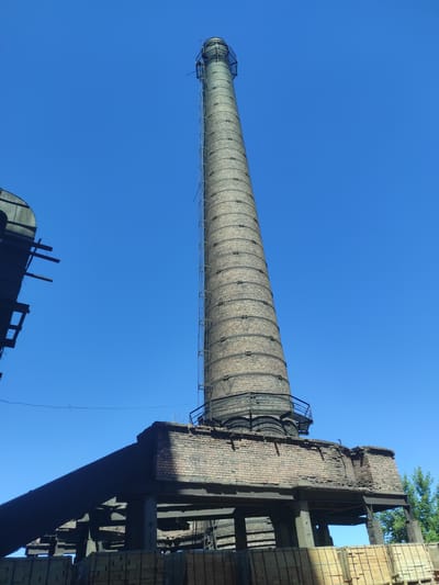 In the city of Kyzyl, Kazakhstan, a tall, weathered brick smokestack rises against a clear, vibrant blue sky. The smokestack, constructed from many courses of reddish-brown bricks, shows signs of age and disuse. A metal ladder, affixed to the exterior, ascends along its entire height, culminating in a caged platform at the very top. The base of the smokestack is anchored to a brick structure with a damaged, crumbling facade, supported by a framework of exposed metal beams and concrete pillars. To the left of the smokestack, a dark, angular structure, possibly part of an industrial building, looms in the foreground, its framework also partially visible. In the lower portion of the image, stacks of rectangular, light-colored objects, possibly bricks or building materials, are neatly arranged. A few green trees are visible in the background on the right, adding a touch of nature to the industrial landscape. The scene appears to be a snapshot of a disused industrial site under a bright, sunny day.