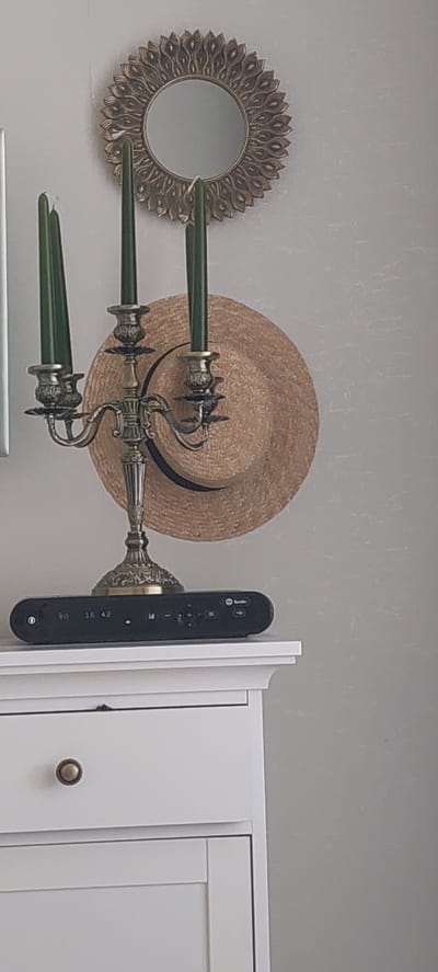 This image depicts a still life arrangement in what appears to be a domestic setting, possibly a dresser or shelf. Dominating the center is an ornate, antique-style brass candelabra holding three dark green tapered candles. To the right of the candelabra, a straw hat with a dark band is casually placed against the wall. Above the candelabra and hat, a decorative, sunburst-style mirror with a metallic gold finish hangs on a textured, pale gray wall. In the foreground, resting on a white surface, is a black electronic device with various buttons and a digital display showing "30 16 42". The electronic device appears to be a remote control or a small entertainment system component. The white surface itself is part of a piece of furniture, possibly a dresser or cabinet, with visible drawer details and a brass-colored round knob. The background wall has a subtle, light texture, possibly wallpaper. There are no people or animals present in the image. The lighting suggests an indoor environment, likely with ambient light from a window or overhead fixture. The scene is static and arranged as a decorative display. There is no information available regarding the location in the city of Ivanovo, Russia.