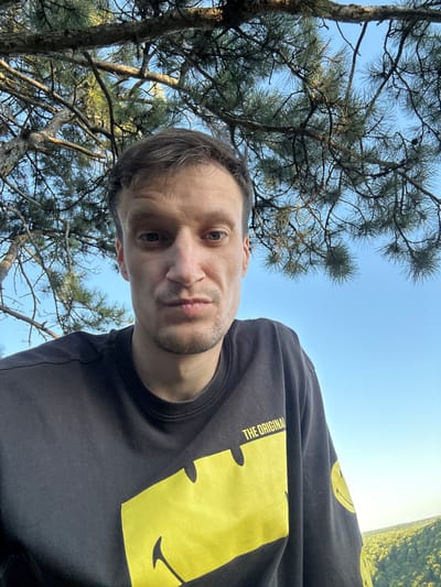 A man is taking a selfie outdoors, looking directly at the camera. He is wearing a black long-sleeved shirt with a yellow smiley face graphic and text that reads "THE ORIGINAL". He has short, dark brown hair and stubble on his chin. Behind him, there are branches and green needles of a conifer tree against a clear blue sky. In the distance, a lush green valley can be seen, suggesting a rural or natural environment, potentially on the outskirts of Pryvorottia Druhe, Ukraine. The lighting suggests it is daytime, possibly late afternoon due to the soft light.