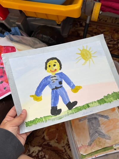 A child's watercolor painting of a person in a blue jumpsuit standing in a field is held up by a hand wearing a gray sweatshirt. The painting depicts a yellow sun in a pale blue sky, with a patch of green grass along the bottom. The figure has a yellow face with black hair, a red mouth, and black eyes. The jumpsuit has black details and a black belt. A name, "Лариса", is written in cursive script on the lower right side of the painting. The painting is placed on top of other children's artwork within a clear plastic container. The background suggests an indoor environment with a patterned rug and part of a yellow and red toy in the upper left corner.