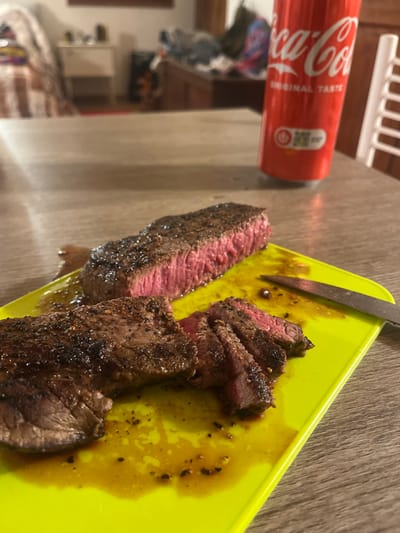 The scene depicts a domestic setting in Monsummano Terme, Italy, focusing on a meal prepared on a light wooden table. In the foreground, a vibrant lime-green plastic cutting board holds two cuts of cooked steak. One long, thick steak piece is severed, displaying a dark, seared exterior crust and a medium-rare, pink interior. Adjacent to it, another steak portion is partially sliced into thinner pieces, revealing a similar pink internal doneness and dark crust. The cutting board is stained with dark brown meat juices and black seasoning particles. A metal knife with a dark handle rests on the right side of the cutting board. Behind the cutting board, a red Coca-Cola can stands upright. The background is blurred but reveals elements of an indoor room: a striped bed or sofa on the far left, a small white dresser, a larger dark wooden dresser with items on top, and a white slatted chair on the far right. No individuals are present.