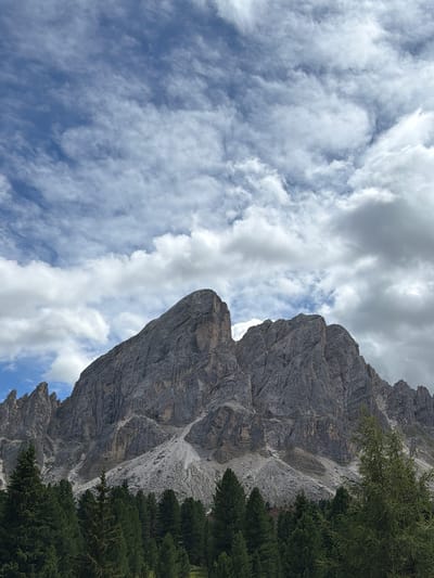 This is a vertical, eye-level, outdoor, full shot of a dramatic rocky mountain face with a bright blue sky filled with fluffy white clouds above and to the left. In the foreground on the right side of the image, there is a lush green conifer tree line. The mountain itself is imposing, with rough, grey, textured rock face that appears to be made of layers. The sky is dynamic with both wispy and dense clouds. The lighting suggests it is daytime, with some areas of the mountain brightly lit and others in shadow, indicating the sun is high in the sky. The scene evokes a sense of grandeur and the power of nature. There is no visible text in the image.