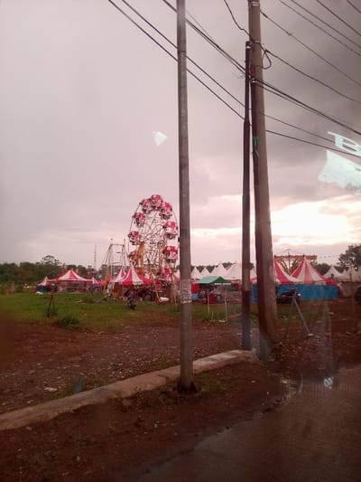 This image captures a temporary fairground or carnival established on an open, unpaved field in Dramaga, Indonesia. The scene is viewed from behind multiple utility poles with overhead electrical wires, suggesting a roadside perspective, possibly through a vehicle window due to visible reflections.
In the foreground, three dark utility poles are prominent, two on the right and one on the left, supporting a network of black electrical wires. The ground directly beneath the poles is dark, wet-looking earth with scattered green vegetation, separated from a paved surface (partially visible lower right) by a low concrete curb.
The midground features the fairground itself. A large, red and white Ferris wheel stands centrally, appearing stationary. Numerous pointed tents, predominantly striped red and white, but also including solid green and blue-and-white variations, are erected across the field. Another metal amusement ride structure, possibly a swing ride, is partially visible to the left of the Ferris wheel. Small clusters of individuals are visible near the tents and rides; one person is positioned on a dark scooter or motorcycle parked on the dirt. The ground within the fair area consists of dark, disturbed earth interspersed with patches of green grass.
The background reveals a dense line of green trees and foliage, behind which a tall metal tower is discernible on the far left. The sky is uniformly overcast, displaying a muted gray coloration with subtle pinkish undertones, suggesting either early morning or late afternoon conditions.