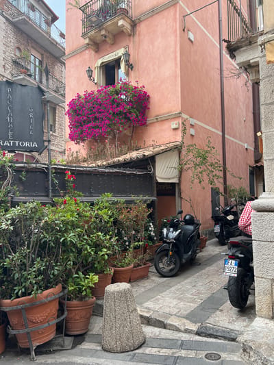 The image depicts a narrow cobblestone alleyway in Taormina, Italy, flanked by buildings with a distinct Mediterranean architectural style. The building on the right is painted a warm, peachy-pink hue and features a balcony adorned with lush pink bougainvillea cascading over its railing. Two black scooters are parked in the alley. The building on the left is constructed of stone and has balconies with ornate iron railings. A dark gray sign with white lettering that reads "RACCANALE GRILL TRATTORIA" is prominently displayed. Greenery, including potted plants and vines, lines the lower section of the alley, creating a vibrant, natural contrast with the man-made structures. The overall atmosphere is one of charming, old-world Italian street life.