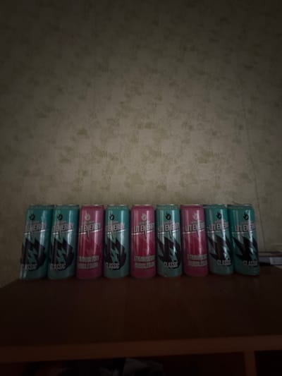 The image displays a still life arrangement of eleven energy drink cans laid out horizontally on a light-colored, subtly patterned floor or surface, possibly linoleum, in Lyman, Ukraine. The cans are primarily of two types: six teal-colored cans and five pink-colored cans, suggesting different flavors. They are arranged in a repeating pattern: two teal, one pink, two teal, one pink, two teal, one pink, and finally two teal cans at the end.

Visible text on the cans identifies the brand as "LITE ENERGY" and the product type as "ENERGY DRINK." The teal cans are labeled "CLASSIC," while the pink cans are labeled "STRAWBERRY BUBBLEGUM." Each can also features a distinctive dark, stylized lightning bolt or wing logo. The lighting is somewhat dim, casting subtle shadows and making the left side of the image darker than the right. The background to the right of the cans is dark and indistinct, possibly a wall or furniture.