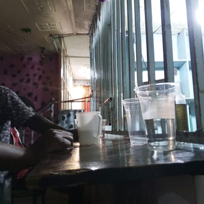 A dimly lit interior scene depicts a table with several glasses and a mug placed on its surface. A person sits at the table, their arms resting on the edge. A barred window is visible behind the table. The ceiling has a patterned design, and the walls appear to be decorated. Based on the prompt, the location is Kinoo ward, Kenya.