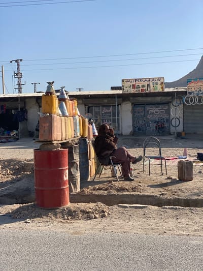 The image displays a street scene in کوکران, Afghanistan, under a clear, bright blue sky. In the foreground, a makeshift fuel station is evident, constructed from two stacked red metal drums serving as a base, topped with a wooden board supporting numerous multi-colored plastic jerry cans. Several metal funnels are positioned on the tops of some jerry cans. To the right, one adult male is seated on a light-colored folding chair, his right leg crossed over his left, with his right hand near his face. He wears dark brown outer clothing and light brown trousers. A silver thermos is visible at his feet. An empty dark-colored metal chair stands adjacent to him, with a brown plastic jerry can and a colorful mat placed on the dusty ground nearby. The background features a row of single-story commercial buildings with corrugated metal roofs. The central building exhibits a large sign advertising "PCO" and mobile phones/electronics in a non-Latin script, while bicycle tires are vertically displayed on the exterior of the adjacent shop to its right. Utility poles and power lines are visible against the sky. A barren mountain range rises in the far background on the right. The ground transitions from a paved road in the immediate foreground to uneven, dirt, and gravel surfaces with shallow trenches leading up to the buildings.