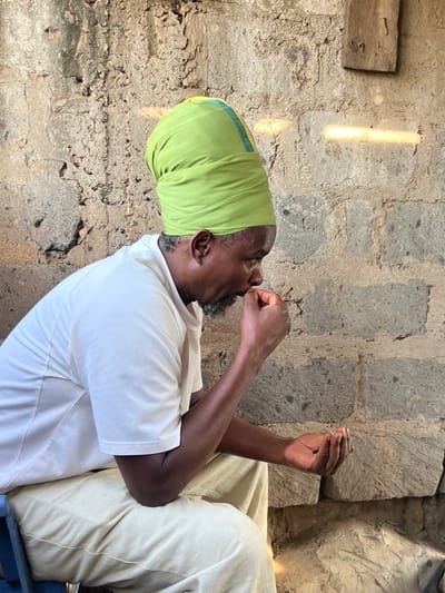 A man wearing a green and yellow head wrap and a white t-shirt is sitting with his back to the camera. His left arm is bent with his hand on his chest and his right arm is bent with his hand in his lap. He is sitting on a blue chair with the back out of view. The scene is in a building with rough, unfinished stone walls. The man is looking down. The scene is in Githurai, Kenya, at the coordinates -1.204 latitude, 36.905 longitude.