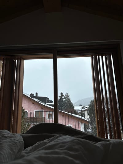 The image shows a view from a window in a room in a building located in Celerina/Schlarigna, Switzerland, at coordinates (latitude: 46.513, longitude: 9.859). The window is open, showing a view of a snow-covered town with several buildings. The closest building is pink with dark windows, a black roof, and a balcony. There is a metal railing on the balcony. Trees are visible in the background. The room contains a bed with white linens, a blanket, and pillows. There is a wooden ceiling above the bed, and the walls are painted a light color. The window is made of wood and has a sliding panel covering half of the window. The window has a paneled covering on the right side. The window has blinds on the right side, all the way up.  The weather appears to be cloudy and cold.