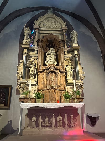 The image depicts an ornate altar in a church, likely in Vianden, Luxembourg. The altar is made of stone and wood, with intricate carvings and sculptures. The central sculpture features a Madonna and Child, surrounded by other religious figures and architectural elements. A white cloth with lace trim drapes over the front of the altar. To the left of the altar is a framed painting. Plants in pots are placed on shelves above the altar, and candles are visible. The floor in front of the altar is illuminated by colored lights, casting a red glow. The background shows a stone archway typical of church architecture.
