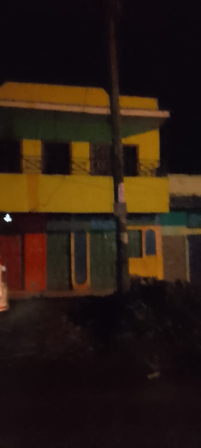 At night, a two-story building is visible. The top floor is yellow with a green awning and a black railing. The bottom floor has several colorful doors or shutters in shades of red, green, and yellow. A utility pole obscures part of the building. The lighting is poor, casting most of the scene into darkness.