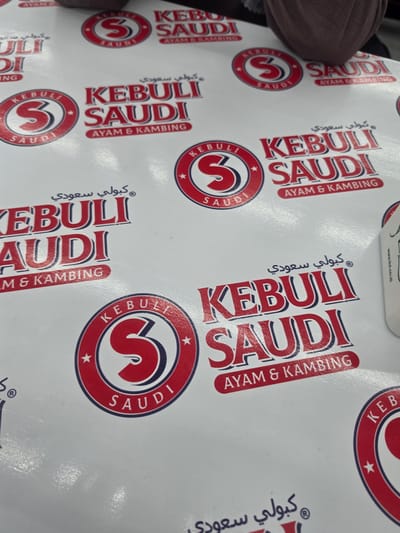 This media file showcases a close-up, overhead view of a white paper tablecloth patterned with repeating logos. The logos feature the brand name "KEBULI SAUDI" in large, stylized red and blue letters, with "AYAM & KAMBING" written below. Each main logo is also encircled by a circular emblem containing the letter "S" within a red and blue border, along with smaller instances of the brand name. Arabic script, reading "كُبولي سعودي" (Kebuli Saudi), is also incorporated into the design.
The tablecloth appears to be laid out on a surface, possibly a table, and is likely intended for food service, given the brand's name suggesting chicken and goat dishes. The lighting suggests an indoor setting, possibly a restaurant or a home dining area. There are no people or specific activities actively taking place within the frame, but the context implies preparation for or engagement in a meal. The image is cropped in a way that suggests the tablecloth might be larger than what is fully visible. The orientation of the tablecloth is slightly askew, as if the photographer is adjusting their position. The text on the tablecloth provides the brand name and type of cuisine, hinting at a Saudi Arabian-inspired dish. There are also some faint and partially visible website addresses at the very top, such as "www.lti.co.id".