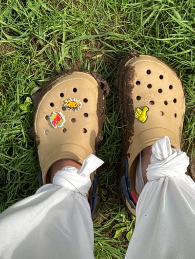 The image shows a view from above of two tan Crocs, covered in mud, on green grass. The Crocs are adorned with novelty charms in the holes. The right Croc has a yellow cartoon hand charm. The left Croc has an "OMG!" and "POW!" comic book-style charm. White pants are tied at the ankles and are tucked into the Crocs. The background consists of green grass, with some patches of dirt and mud. The location of Nozhovka, Russia, is not relevant as it is not visible in the image.