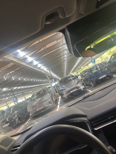 Here is a description of the image:
The image is taken from inside a car, looking out at a parking area in Sint-Niklaas, Belgium. The view through the windshield shows rows of parked vehicles under a metal structure that provides shade. Number plates are visible on some of the cars, with Belgian license plate formats "2-HVT-864", "2-HSP-577", "2-HGD-802" and "2-HDE-413". Parking signs with a "P" are also visible along the lanes. The interior of the car is visible, including the steering wheel and parts of the dashboard. The image seems to be taken during daylight.