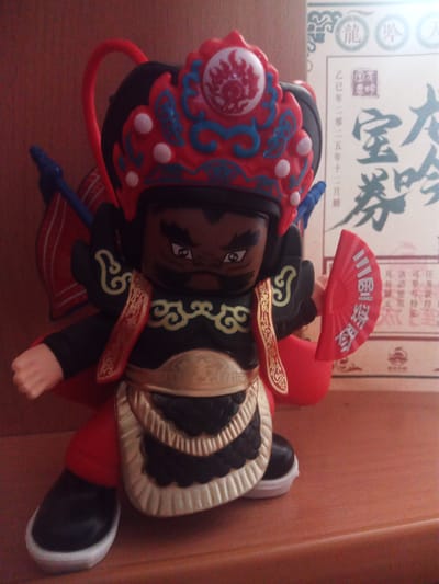 The image shows a figurine of a character dressed in traditional Chinese opera attire. The figurine is standing on a wooden surface, likely a table or shelf, with a blurred background that appears to be a wall and a paper document. The figurine is wearing a red and blue headdress with white circular accents, a black garment with gold trim and red accents, and red pants. It holds a small red fan in its right hand, which has Chinese characters printed on it. The figurine has a dark complexion and a stern expression. The paper document in the background has Chinese characters and some circular seals. The overall lighting suggests it's an indoor setting, likely a room. There is no identifiable information in the image that would allow for the localization of this scene in Usman, Russia.