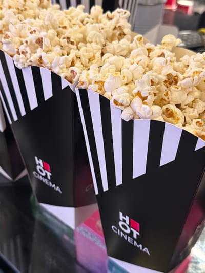 The image shows two black and white striped popcorn containers filled with popcorn. The containers have the words "HOT CINEMA" printed on them. The popcorn is overflowing from the containers. The scene appears to be indoors, possibly at a cinema in Rehovot, Israel, given the name "HOT CINEMA."
