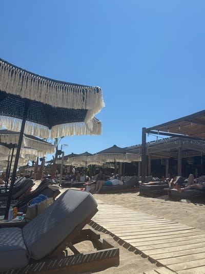 The image depicts a sunny day at a beach resort in the city of Pallini Municipal Unit, Greece. In the foreground, several empty lounge chairs are set up on a sandy beach with a wooden walkway. These chairs are made of dark-colored fabric with wooden frames and are arranged under large black umbrellas with white fringe detailing. In the background, more lounge chairs and umbrellas are visible, with several people relaxing on them. Further back, there are open-air structures with thatched roofs and string lights, suggesting a dining or bar area. Palm trees are scattered throughout the scene, and the sky is a clear, bright blue. The overall atmosphere is one of a relaxed beach vacation.