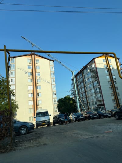 The image depicts an urban scene in Durlești, Moldova, with two multi-story apartment buildings dominating the background. A construction crane is visible in the sky, extending over the buildings. In the foreground, several vehicles are parked, including a white van with "103" displayed on its windshield, a dark sedan, and other cars. Two individuals are standing near the vehicles, appearing to interact. The ground is a mix of paved surfaces and dirt. The sky is clear and blue, suggesting a daytime scene. Trees are present on the left and right sides of the frame, bordering the buildings. A yellow pipe structure runs across the upper portion of the image, obscuring parts of the sky and crane.