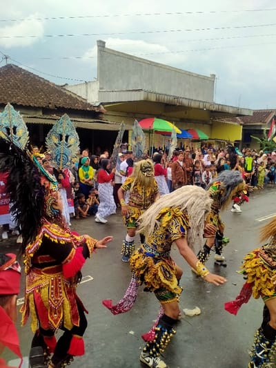 The image depicts an outdoor cultural performance or parade occurring on a wet asphalt street in Mojogedang, Indonesia, under an overcast sky. In the foreground, multiple individuals are actively participating. On the left, a performer is visible from the back, adorned in an elaborate red, gold, and black costume with a large, dark feathered headdress and ornate shoulder pieces. Their body is angled to the right, arms slightly raised. To their right, at least three individuals are dressed in intricate gold, green, and black patterned costumes, each featuring a large, imposing mask with long, shaggy hair, characteristic of a "Barongan" or similar traditional performance. These masked performers have decorative ankle coverings with numerous spherical bells and are captured in mid-movement, suggesting a dance.
Behind these primary performers, several other individuals carry large, ornate, fan-like objects with blue and gold patterns. A dense crowd of spectators, including adults and children, lines both sides of the street, observing the event. Some individuals in the crowd are seated on the ground. In the background, various buildings are visible, including older structures with brown tiled roofs and a more modern-looking building with a flat, light-colored facade. Several colorful open umbrellas (red, green, blue) are present within the spectator area. Overhead electrical wires traverse the sky. A crumpled white object lies on the street surface near the masked performers. A red and white flag is partially visible on the far right.