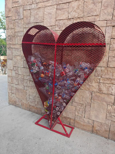 A red, heart-shaped metal mesh bin filled with plastic bottles and aluminum cans is positioned against a beige stone-tiled wall. The bin is supported by a red metal frame. A concrete sidewalk runs in front of the bin and the wall. The scene appears to be outdoors, possibly in a public area or park, with some vegetation visible in the background. The overall impression is that of a recycling station. The location in Cancún, Mexico, is not directly discernible from the image itself, but the presence of tropical foliage might suggest a warm climate.