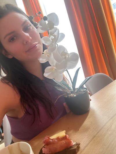 The image shows a close-up selfie of a woman holding a potted orchid with white flowers. She is wearing a purple ribbed tank top and has long, dark hair. The background features orange curtains and a bright window. A small piece of food, possibly toast with toppings, is visible on a wooden table in the foreground, along with a white mug. The lighting suggests an indoor setting, likely a home or cafe, and the presence of an orchid and curtains implies a domestic environment. No specific details about the city of Novoselye, Russia are discernible from the image.