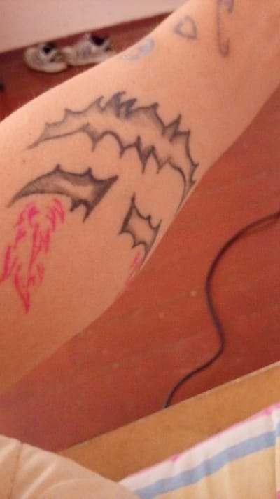The image shows a close-up view of a person's arm with a tattoo of a dragon. The dragon is depicted in black ink with some pink accents. In the background, a pair of small, white sneakers are visible, placed on a reddish-brown surface. The scene appears to be indoors, possibly a bedroom, given the presence of a striped fabric in the lower right corner. There are no discernible landmarks or clues that would indicate the location in Tomsk, Russia.