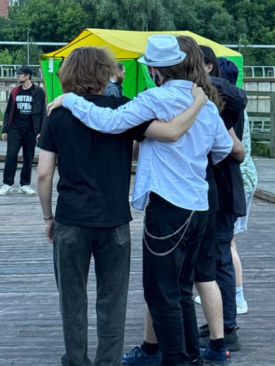 The image depicts a group of five individuals standing on a wooden boardwalk. The individuals are primarily seen from the back, with their faces obscured or turned away.
In the foreground, two individuals are prominently featured, with one person's arm draped around the other's shoulders. The person on the left wears a black t-shirt and dark, possibly denim, pants. The person on the right wears a white collared shirt, black pants adorned with a silver chain, and a white fedora. Their postures suggest an embrace or close companionship.
Behind them, three other individuals are visible. One person, to the far left, wears a black baseball cap, glasses, and a black jacket over a black t-shirt with Russian text. They are looking to the left. To the right of this individual, another person is partially visible, wearing dark clothing. The furthest individual on the right wears a patterned shirt and light-colored shorts or skirt.
The environment appears to be outdoors, with a green and yellow tent structure visible in the background. Trees and foliage are also present, suggesting a park or natural setting. The boardwalk is made of weathered wooden planks.
The presence of Russian text on the t-shirt of the individual on the far left may suggest a connection to Russia, but a definitive identification of the location as Votkinsk, Russia cannot be made solely from the visual information provided. The scene appears to be a casual gathering of young people.