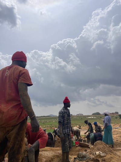 The image depicts a group of people working in Maiyanga, Nigeria. The workers are covered in mud. Some are digging with shovels. Some are standing and observing. There is vegetation and livestock in the background. The sky is cloudy.